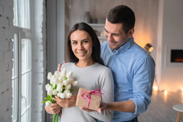 Tiro de alegre joven caucásico que dando caja regalo a su esposa con mirada en ella. Hermosa mujer con flores en las manos en el interior del hogar y mirar en la cámara, feliz pareja — Foto de Stock