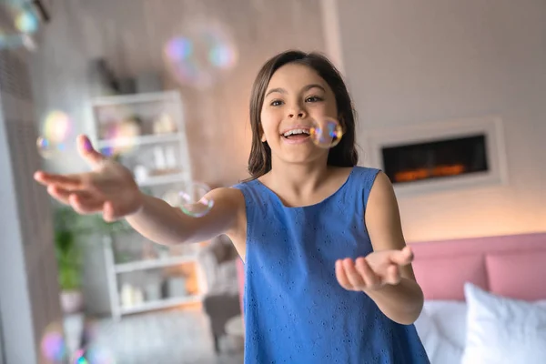 Close up tiro de menina feliz brincando com o ventilador de bolhas em casa perto da cama com lareira no fundo — Fotografia de Stock