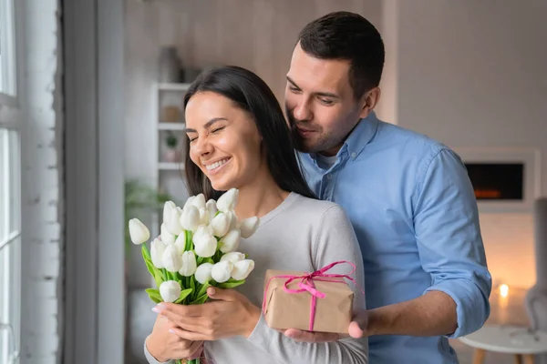 Κοντινό πλάνο του εύθυμου νεαρού καυκάσιου άντρα που δίνει το παρόν κουτί στη γυναίκα του με το βλέμμα του. Όμορφη γυναίκα ενθουσιασμένος με τα λουλούδια στα χέρια στο σπίτι εσωτερικό, ευτυχισμένο ζευγάρι — Φωτογραφία Αρχείου