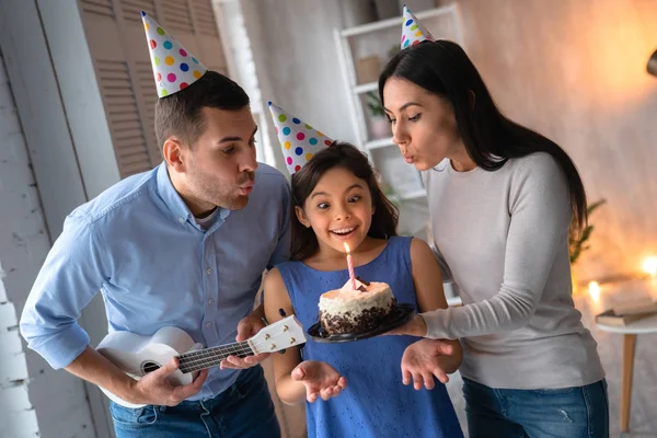 Joven padre y madre celebrando el cumpleaños de su hija. Familia vistiendo sombreros de fiesta y celebrando el cumpleaños de su hija en casa. Pequeña linda chica soplando vela en pastel de cumpleaños mientras su fa — Foto de Stock