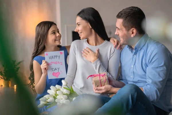 Lycklig mors dag! Framifrån bild av lycklig familj firar Mors dag. Ung man ger en gåva hans hustru som sitter på golvet med blommor i händerna. Liten flicka med nuvarande kort för mamma — Stockfoto