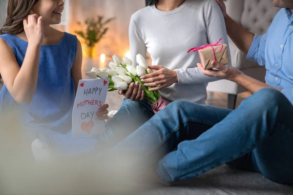 Lycklig mors dag! Närbild skott av lycklig familj firar Mors dag. Ung man ger en gåva hans hustru som sitter på golvet med blommor i händerna. Liten flicka med nuvarande kort för mamma — Stockfoto