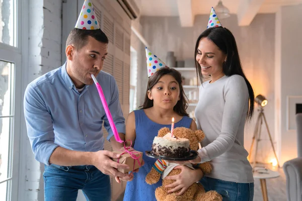 Ung far och mor firar sin dotters födelsedag. Familjen bär partyhattar och firar dotters födelsedag hemma. Liten söt flicka blåser ut ljus på födelsedagstårta — Stockfoto