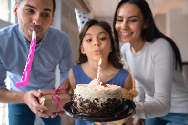 Молодые отец и мать празднуют день рождения дочери. Семья в шляпах для вечеринок и празднует день рождения дочери дома. Маленькая милая девочка задувает свечи на торте на день рождения — стоковое фото