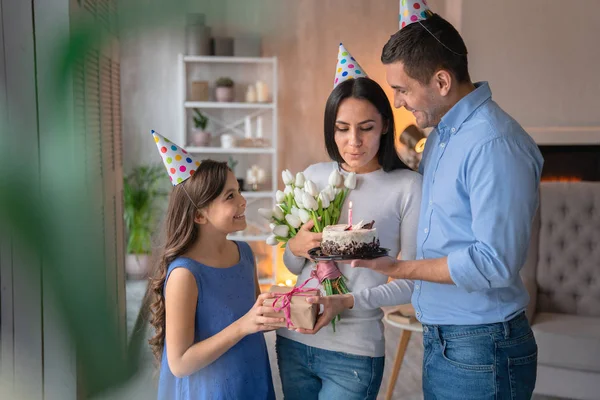 Feliz familia celebrando el cumpleaños de mamá. Hombre de pie con pastel de vacaciones en la mano, niña con caja de regalo para su madre con flores en las manos —  Fotos de Stock