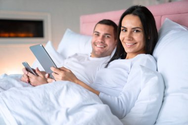 Arka planda kamera ve şömine bakmak ile elinde tablet ve cep telefonu ile yatakta yatan mutlu genç çift