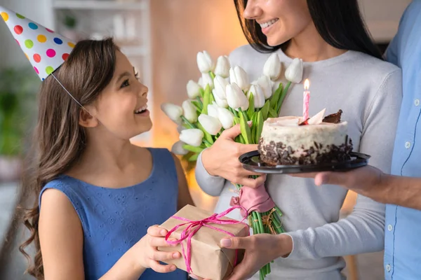 Jovem pai e mãe comemorando o aniversário de sua filha. Família vestindo chapéus de festa e celebrando o aniversário da filha com bolo de aniversário e presente em casa — Fotografia de Stock