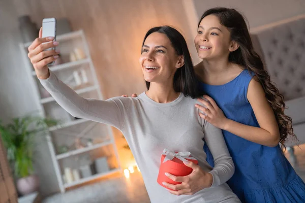 Menina bonito feliz parabenizar sua mãe. Mulher caucasiana morena alegre com presente no aniversário de sua filha em suas mãos fazendo selfie com sua filha. Feliz dia da mãe ! — Fotografia de Stock