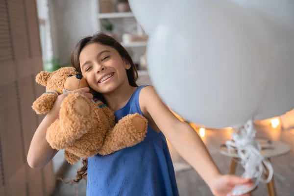 Feliz linda niña abrazando a su osito de peluche con globos de aire en su mano —  Fotos de Stock