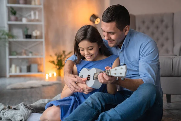 Menina sorridente bonito tocando na guitarra ukulele com seu pai. Pai feliz ensinando a tocar no ukulele guitarra sua filha em casa — Fotografia de Stock