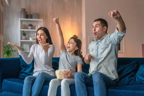 Emocionales padres jóvenes y su linda hijita están viendo películas en la televisión, comiendo palomitas de maíz y sentados en el sofá en casa — Foto de Stock