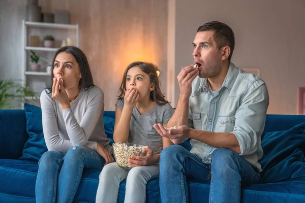 Emocionados padres jóvenes y su linda hijita están viendo películas en la televisión, comiendo palomitas de maíz y sentados en el sofá en casa — Foto de Stock