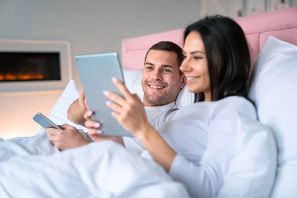 Mujer alegre mostrando algo en tableta a su marido. Sonriente hombre acostado en la cama con su esposa sosteniendo el teléfono móvil en sus manos con enfoque en el hombre —  Fotos de Stock