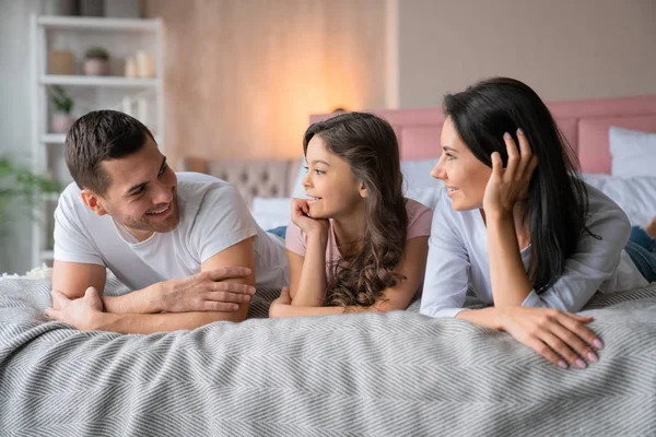 Munter kaukasisk mand og hans smukke lille datter kigger på hinanden, mens de ligger i sengen med smilende mor - Stock-foto