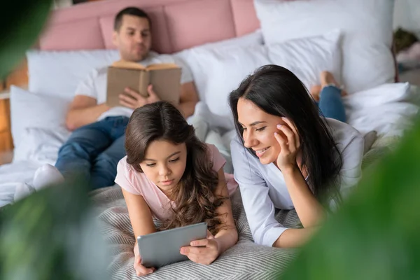 Linda niña mostrando algo a su madre en la tableta mientras está acostada en la cama con el padre que la lectura de libro en el fondo —  Fotos de Stock