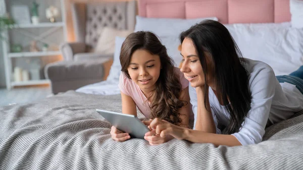 Hermosa mujer joven mostrando algo a su linda hija sonriente en la tableta en la cama —  Fotos de Stock