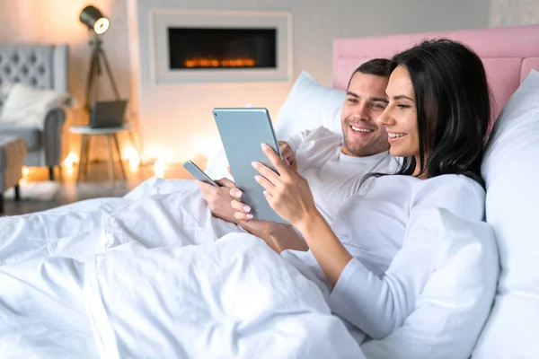 Mujer sonriente mostrando algo en tableta a su marido. Sonriente hombre acostado en la cama con su esposa sosteniendo el teléfono móvil en sus manos — Foto de Stock