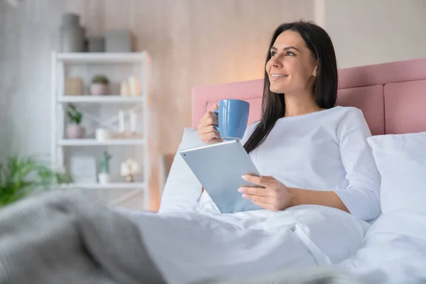 Momen mimpi. Tampilan samping foto wanita cantik muda dengan piyama di ranjang rumah dengan secangkir kopi pagi dan tablet di tangan — Stok Foto