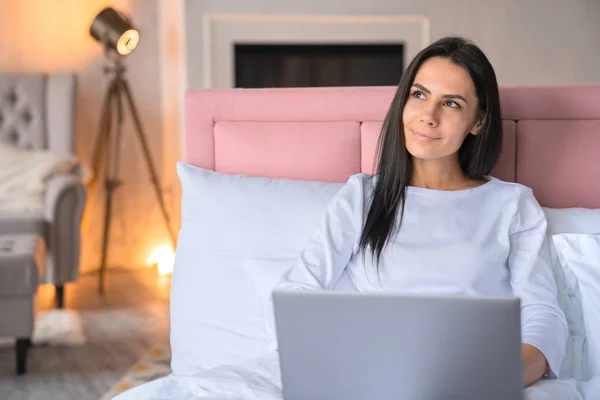 Sonriendo hermosa mujer usando portátil mientras está acostado en la cama en casa con soñando mirar hacia otro lado y lámpara de pie en el fondo —  Fotos de Stock