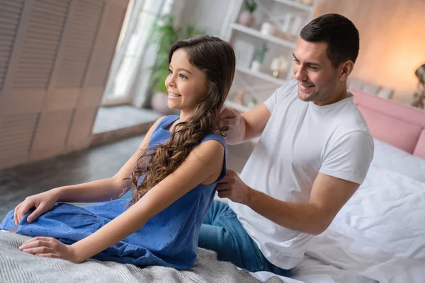 Sonriente padre peinando el pelo a su pequeña hija linda en vestido sentado en la cama en casa — Foto de Stock