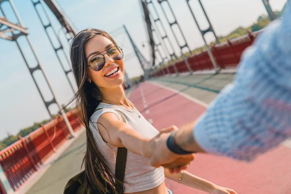 За мной! Счастливая молодая женщина тянет парня за руку, рука об руку, идя в яркий солнечный день по мосту — стоковое фото