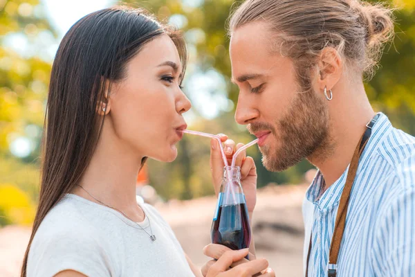 Unga lyckliga par utomhus dricker kall dryck från glas flaska med halm. Side View of man och kvinna dricker läsk och leende. — Stockfoto