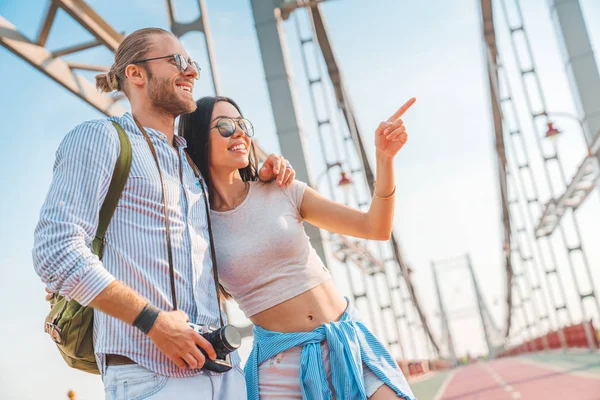 Feliz pareja exploradora de pie al aire libre. Hombre hablando con la mujer con su brazo alrededor de sus hombros y ella señalando hacia — Foto de Stock