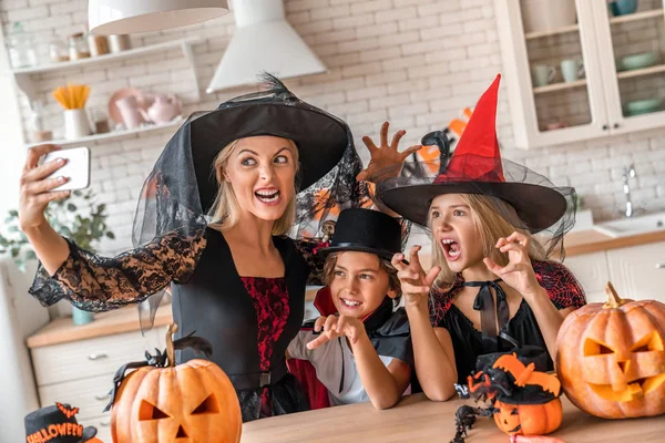 Uśmiechnięta młoda kobieta robi sobie selfie i bawi się z dziećmi w urządzonej kuchni Halloween — Zdjęcie stockowe