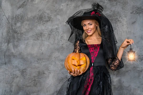 Imagem da jovem mulher feliz vestida com traje de bruxa halloween sobre fundo cinza com abóbora e lanterna — Fotografia de Stock