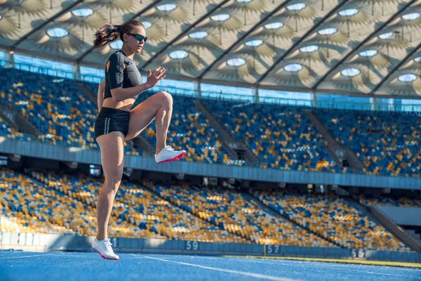 Young woman runner athlete stretching legs before run during sunny morning on stadium track — Stock Photo, Image