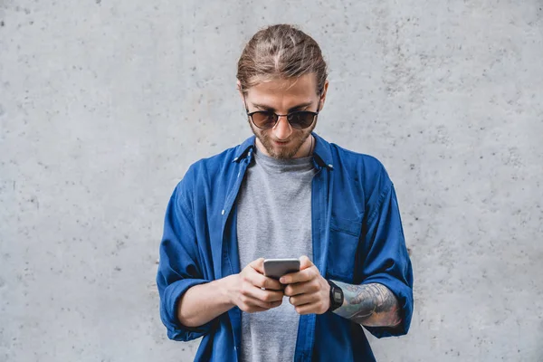 Jovem mensagem de texto no telefone inteligente isolado em fundo cinza. Sorrindo caucasiano masculino em óculos de sol segurando smartphone e olhando para ele . — Fotografia de Stock