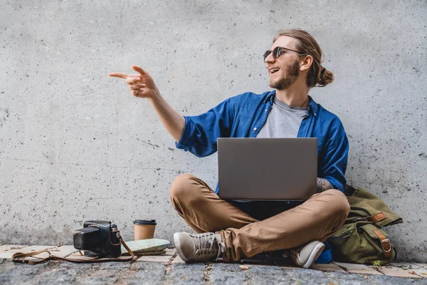 Jovem sorridente sorrindo homem olhando para longe e encontrar alguém ao usar laptop ao ar livre. O conceito de estudo, freelancer, trabalho, tecnologia, viagens — Fotografia de Stock