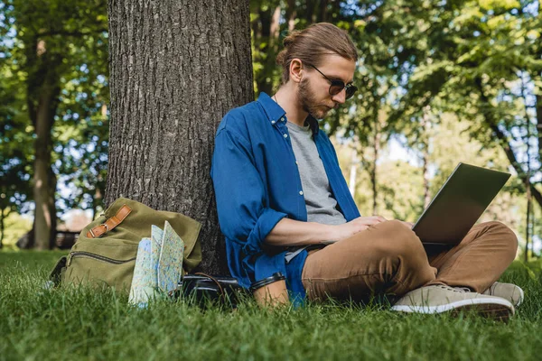 Homem usando óculos de sol usando laptop enquanto sentado debaixo de uma árvore no parque. Jovem sentado na grama no parque trabalhando no laptop enquanto viaja — Fotografia de Stock