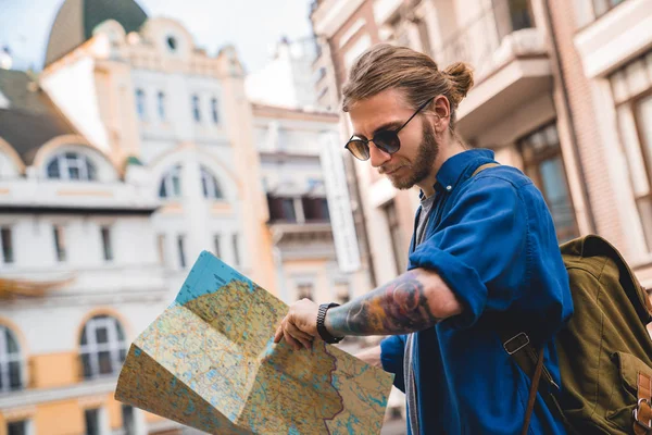 Guapo joven en ropa casual sosteniendo mapa y comprobando la hora mientras está parado en las calles de la ciudad al aire libre — Foto de Stock