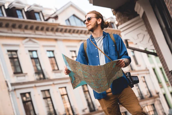 Handsome young man in casual clothing holding map and looking away while standing on the city street outdoors