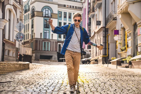 In voller Länge wandert ein junger Mann in legerer Kleidung durch die leere Straße und amüsiert sich beim morgendlichen Kaffee im Freien. Sommerreise. — Stockfoto