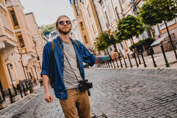 Schöner junger Mann mit Fotokamera in lässiger Kleidung, der wegschaut, während er draußen steht und versucht, ein Auto anzuhalten — Stockfoto