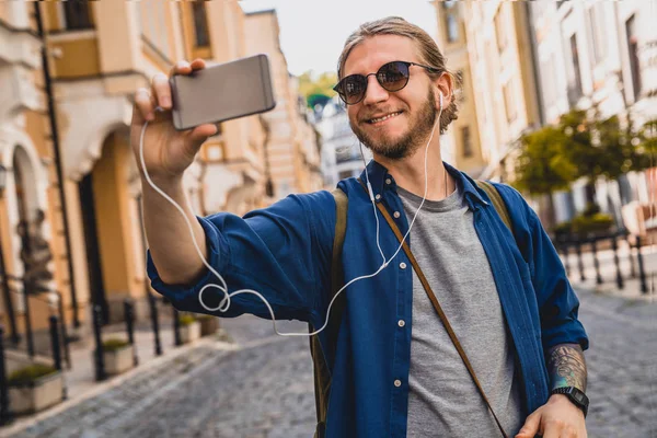 Pemuda tampan dengan pakaian kasual tersenyum sambil selfie di luar ruangan. turis Kaukasia berbicara dengan teman-teman melalui smartphone saat berjalan di jalan-jalan kota Stok Foto