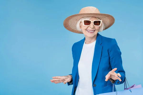 资深妇女购物者戴着太阳镜 头戴安全帽 头戴蓝色背景的购物袋 — 图库照片