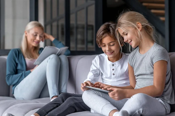 Çocuklar dijital tablet kullanırken anneler oturma odasındaki kanepede kitap okuyor.