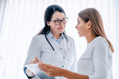 Gülümseyen kadın doktor sağlık raporunu tutuyor ve kadın hastayla konuşuyor.