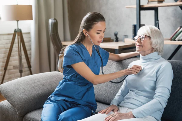 Medical assistance. Asian nurse using stethoscope for elderly woman indoors