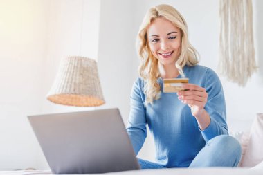 Yatakta dizüstü bilgisayarla otururken online alışveriş için kredi kartı kullanan mutlu beyaz kadın portresi.