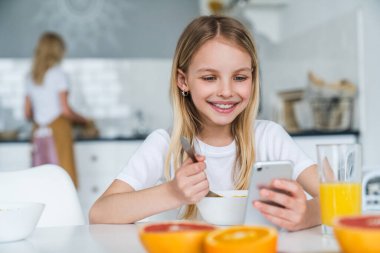 Küçük kız evde telefon kullanırken kahvaltı yapıyor.