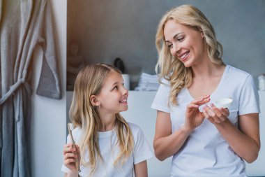 Mother and daughter child girl are brushing teeth toothbrushes and using skin cream in the bathroom clipart