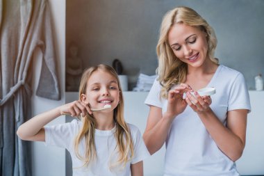 Little girl brushing teeth while her mother using cream in the bathroom clipart