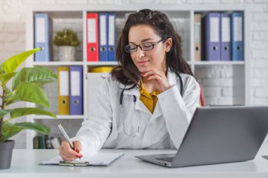 Dizüstü bilgisayarda çalışan genç bayan doktor iş yerinde not alıyor.