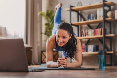 Formda bir kadın evdeki oturma odasında antrenman yaparken dizüstü bilgisayar başında yoga yapıyor ve özel ders seyrediyor.