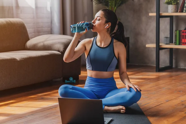 穿着运动服的女人坐在垫子上 喝着一瓶水 同时在家里客厅里使用笔记本电脑 — 图库照片