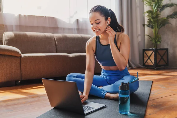 Spor ve eğlence konsepti. Spor giyimli ve kulaklıklı bir kadın oturma odasında dizüstü bilgisayar kullanıyor..